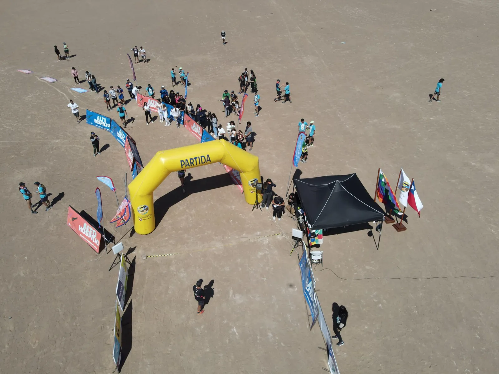 Toma aérea con drone de un arco amarillo, una carpa, banderas de marcas y personas sobre la arena del desierto